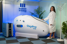 Load image into Gallery viewer, Woman in a bathrobe standing next to an OxyPro hyperbaric chamber in a Recovery Studio.